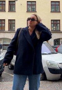 Marineblauer Wollmantel mit doppelt geknöpftem Design, breiten Revers, strukturiertem Stoff und übergroßen Ärmeln, kombiniert mit hellblauen Jeans und Sonnenbrille.