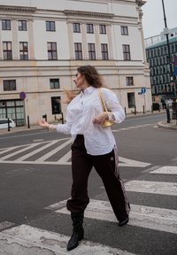 Camisa blanca abotonada sobre pantalones deportivos burdeos oscuros con rayas blancas y logo; botas negras hasta la rodilla con tacón; pequeño bolso amarillo.