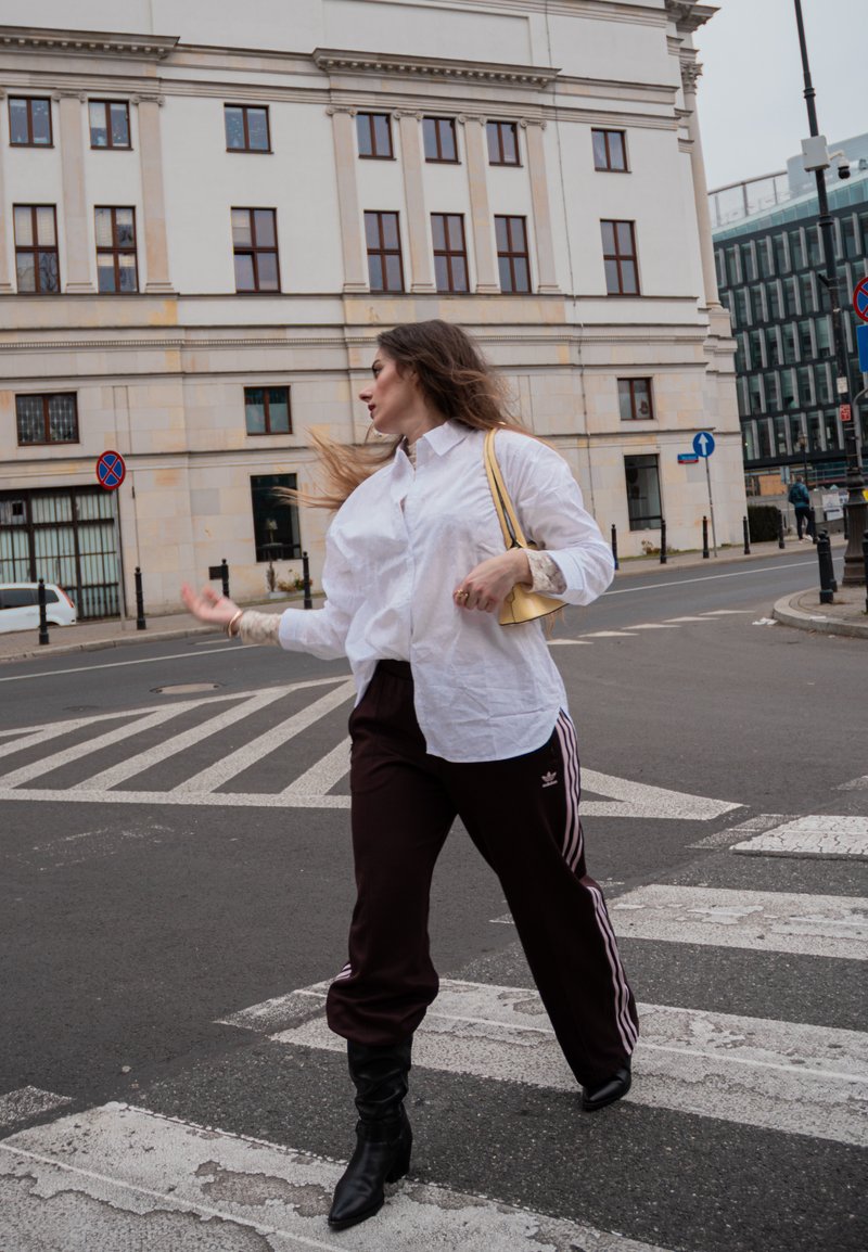 Chemise blanche boutonnée portée sur un pantalon de survêtement bordeaux foncé avec des rayures blanches et un logo ; bottes à talons hautes noires jusqu'aux genoux ; petit sac à main jaune.