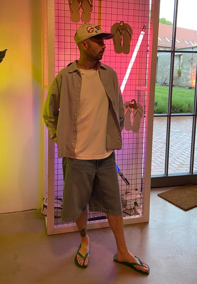Hombre con gorra, camisa de rayas, camiseta blanca, pantalones cortos vaqueros largos y chanclas, de pie en el interior junto a una exposición de chanclas con luces de neón.