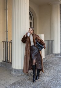 Manteau en fausse fourrure marron avec de gros boutons noirs, pull en maille crème, jupe en cuir marron fendue, bottes noires montantes à talons, et sac noir.