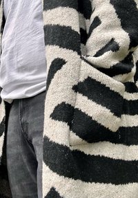 Pletený kardigán s čierno-krémovým zigzag vzorom, s otvoreným predným dielom a mäkkou textúrou, nosený cez biele tričko a tmavé džínsy.