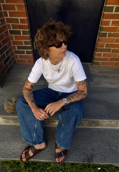 Persona sentada en las escaleras, vistiendo una camiseta blanca, jeans anchos azul oscuro y sandalias marrones. Tatuajes notables y joyería de plata en capas.