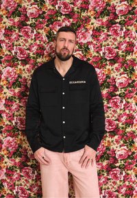 Camisa de botão preta de mangas longas com dois bolsos no peito e detalhes em botão branco, usada com calças rosa claro contra um fundo floral.