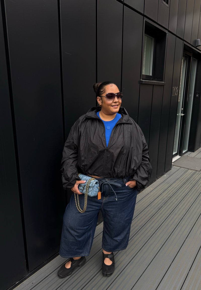 Black cropped jacket, blue shirt, wide denim culottes, black slip-on shoes, and a denim pouch with a chain. Sunglasses complete the look.