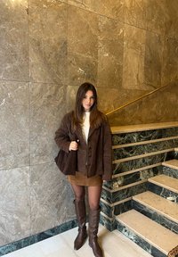 Femme en veste marron, jupe et bottes, portant un sac assorti marron, debout près d'un mur en marbre et d'un escalier avec une rampe dorée.