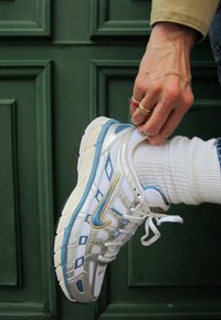 Scarpa sportiva bianca con accenti blu e oro, parte superiore in rete, suola testurizzata e lacci. Indossata con una calza bianca su uno sfondo verde.