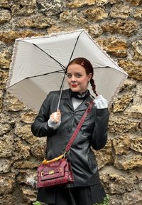 Czarna skórzana kurtka, szary sweter, czarna spódnica, trzyma białą parasolkę z falbanką oraz czerwoną torebkę w wzory ze złotymi elementami.