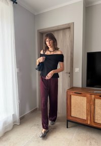 Sieviete melnā topā ar plikiem pleciem, bordo biksēs, čūskas raksta apavos, rokās turot melnu somu, stāv gaišā istabā pie koka skapja un aizvērtām durvīm.