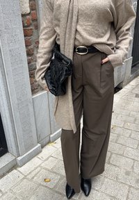 Person in beigem Pullover und braunen, weiten Hose mit schwarzem Gürtel, hält eine schwarze geflochtene Clutch und steht auf einem gepflasterten Bürgersteig.