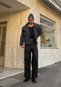Homme portant un pantalon large noir, une veste sombre, des lunettes de soleil orange et un bonnet gris, debout devant un salon avec un panneau "Cut and Brush".