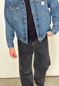Denimska jakna v svetlo modri barvi z dvema žepoma na prsih in gumbi, v kombinaciji z črnimi širokimi hlačami. Gladka tekstura, sproščen kroj.