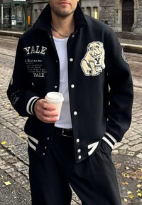 Zwarte varsity-jack met een bulldogafbeelding, Yale- borduursel, witte strepen op de mouwen en contrasterende knopen. Houdt een koffiekop vast.