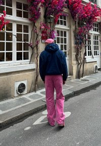 Άτομο που φοράει μπλε κουκούλα και ροζ φαρδι παντελόνι, περπατώντας σε έναν δρόμο με μωβ ανθοστολίσματα και λευκά παράθυρα.