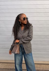 Γυναίκα με μακριά σγουρά μαλλιά που φοράει γυαλιά ηλίου, γκρι σακάκι, μπλε τζιν και κρατάει ένα καφέ καρό τσάντα μπροστά σε γκρι τοίχο.