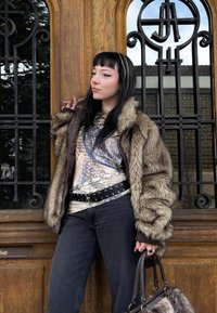 Giacca marrone in pelliccia sintetica con maniche testurizzate, camicia a fantasia, jeans neri, cintura argentata e borsa scura, ambientati davanti a una porta di legno.