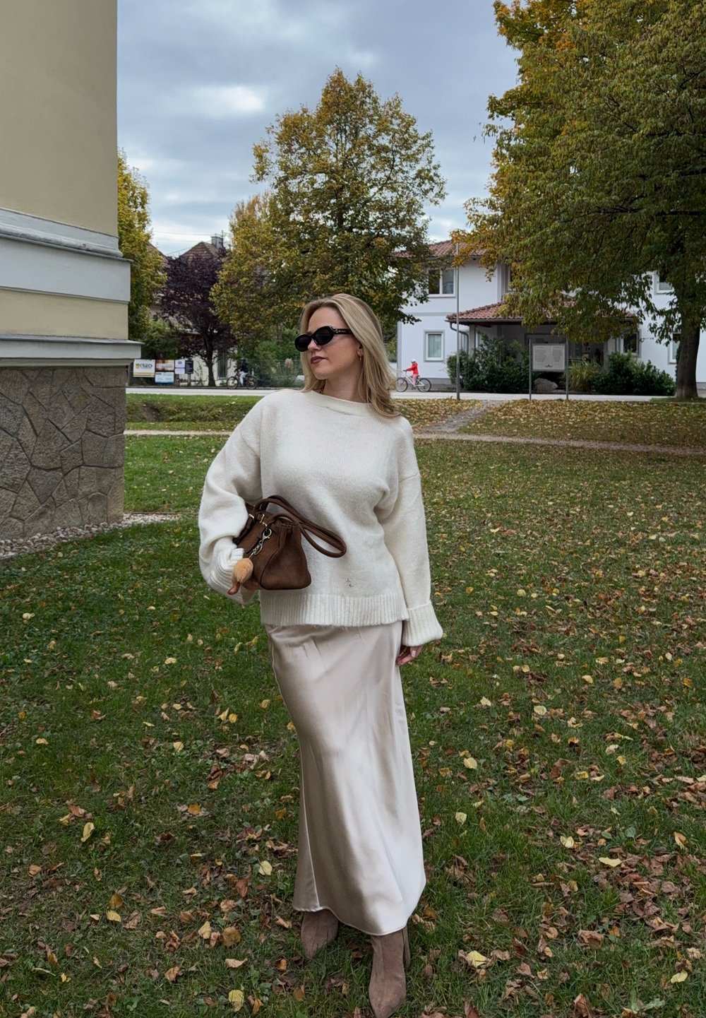 Pull en tricot crème à coupe ample, associé à une jupe beige en satin et un sac à main brun. Le mannequin porte des lunettes de soleil noires et se tient sur de l'herbe.