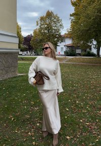 Pull en tricot crème à coupe ample, associé à une jupe beige en satin et un sac à main brun. Le mannequin porte des lunettes de soleil noires et se tient sur de l'herbe.