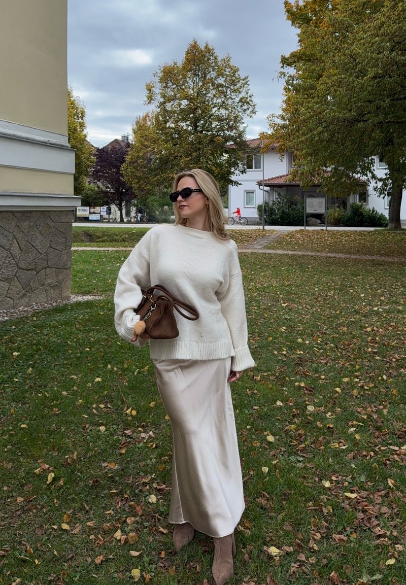 Maglione lavorato a maglia color crema con vestibilità morbida, abbinato a una gonna satinata beige e a una borsa marrone. Il modello indossa occhiali da sole neri, in piedi sull'erba.