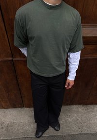 Personne portant une chemise à manches courtes vert olive superposée sur une chemise blanche à manches longues, un pantalon noir et des chaussures noires, debout contre un mur en bois.