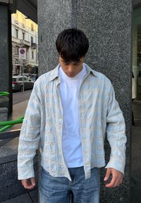 Koszula w drobną kratę w jasnoniebieskim i kremowym kolorze, z teksturowanego materiału, z rozpiętym kołnierzem, noszona na białym t-shircie i zestawiona z niebieskimi dżinsami.