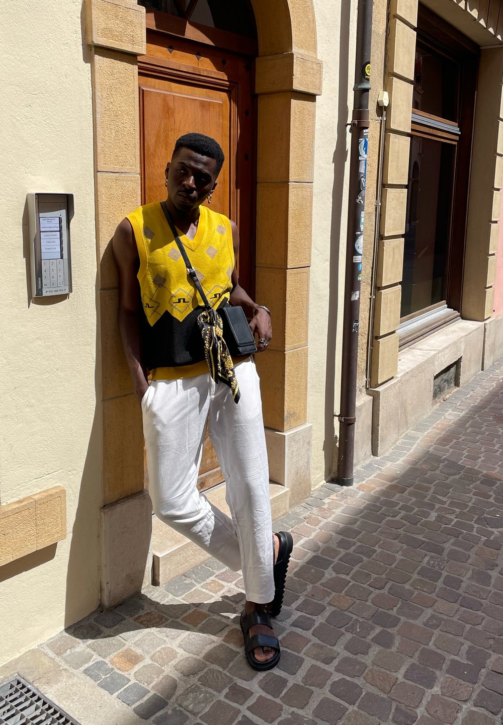 Κίτρινο και μαύρο καπιτονέ αμάνικο πουλόβερ, λευκά λινά παντελόνια, μαύρη τσάντα χιαστί, μαύρες παντόφλες, όρθια μπροστά σε έναν πέτρινο τοίχο.