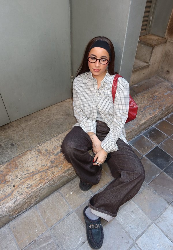 Noor naine prillide, peavõru, ruudulise särgi ja lahtiste pükstega istub kivist trepil, hoides käes musta eset, mille õlal on punane kott.