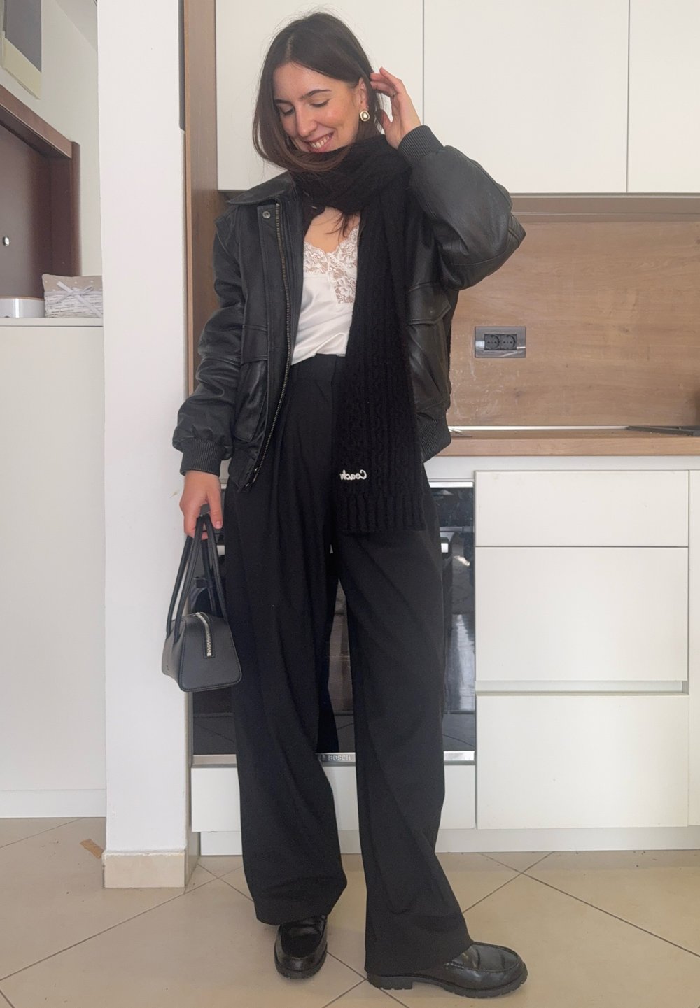 Femme souriante touchant ses cheveux, portant une veste en cuir noire, une écharpe noire, un haut blanc en dentelle, un pantalon large noir, tenant un petit sac à main noir.