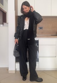 Femme souriante touchant ses cheveux, portant une veste en cuir noire, une écharpe noire, un haut blanc en dentelle, un pantalon large noir, tenant un petit sac à main noir.