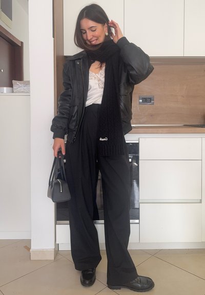 Mujer sonriendo y tocándose el cabello, lleva chaqueta de cuero negra, bufanda negra, blusa blanca de encaje, pantalones anchos negros y sostiene un pequeño bolso negro.
