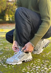 Personne portant un pull vert et un jean foncé qui lace des baskets blanches Nike en plein air sur un chemin de pierres couvert de mousse.