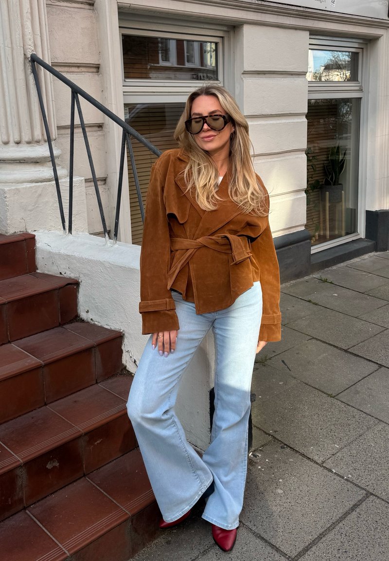 Γυναίκα με μακριά ξανθά μαλλιά που φοράει μεγάλα σκούρα γυαλιά ηλίου, καφέ μπουφάν με ζώνη, ανοιχτά μπλε καμπάνα τζιν και κόκκινα μυτερά παπούτσια, στέκεται δίπλα στις σκάλες.