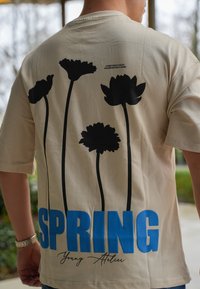 Beige kortärmad t-shirt med svarta blommönster och "SPRING" i fetblå text längst ner. Inkluderar varumärket "Young Atelier."