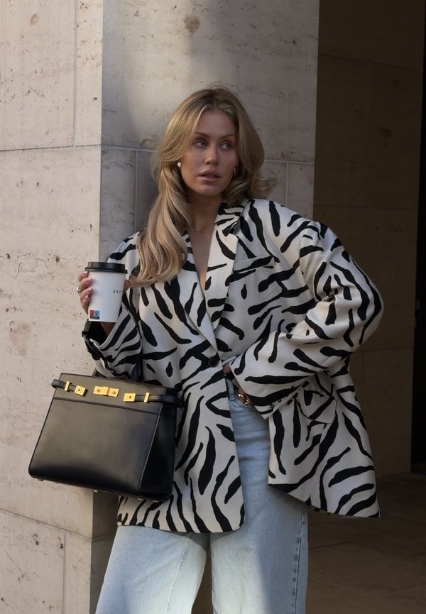 Mujer con una chaqueta con estampado de cebra en blanco y negro, sosteniendo una taza de café y un bolso negro, de pie junto a una pared de piedra beige.
