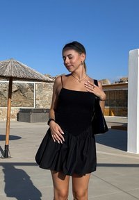 Črna sončna obleka z oprijetim, smokiranim zgornjim delom in razširjenim krilom. Ima tanke naramnice in teksturirano tkanino, postavljeno na sončno ozadje.