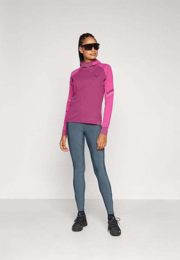 Sportovní oděv zahrnující růžové tričko s dlouhým rukávem a vysokým límcem, doplněné šedými legínami. Materiál vypadá hladce a má obepínající střih. Nosí se černé sluneční brýle.