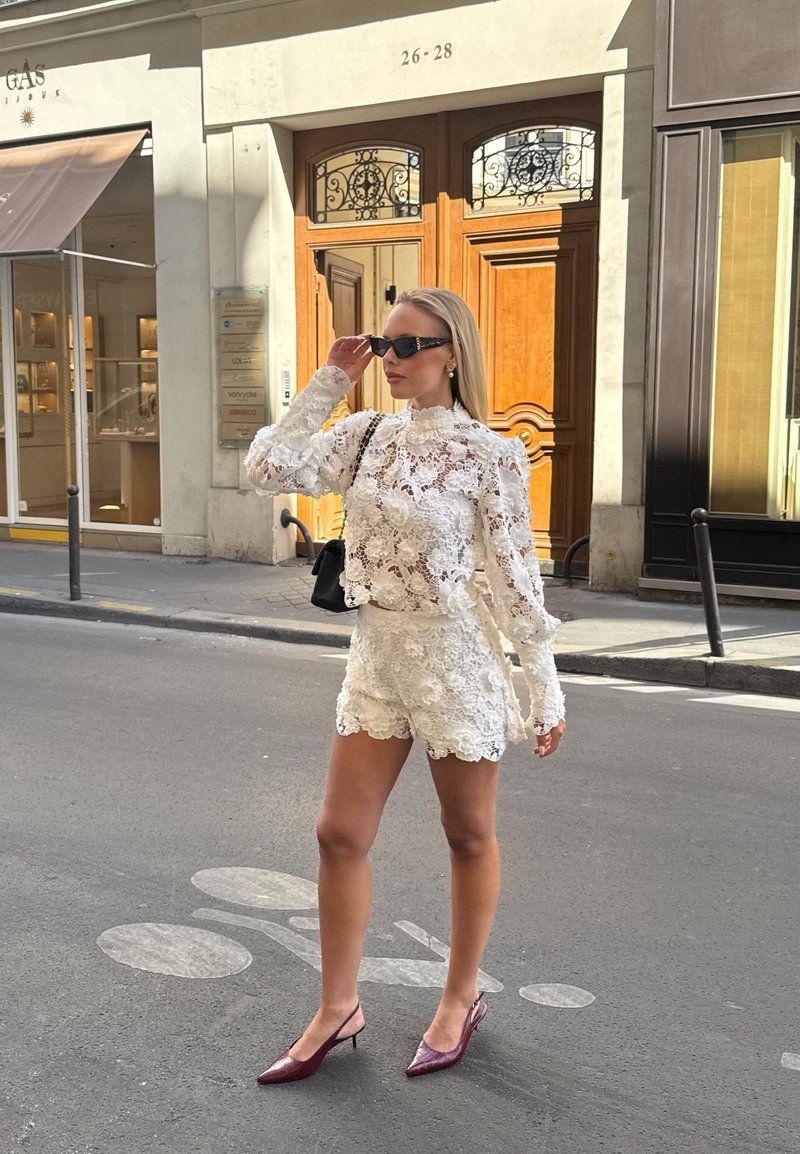 Une femme ajuste des lunettes de soleil noires, porte un chemisier en dentelle blanche et un short avec des talons bourgogne, se tenant dans une rue de la ville près de portes en bois.