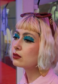 Junge Person mit blonden Bob-Haar, auffälligem blauen, gewellten Lidschatten, rotem Lippenstift, Septum- und Lippenpiercings, großer goldener Ohrringe und roten Sonnenbrillen auf dem Kopf.