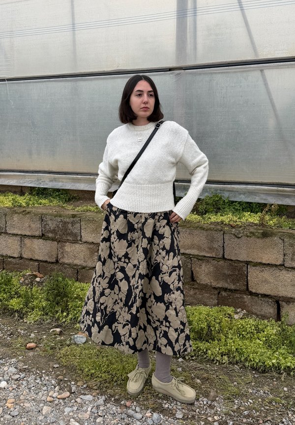 Femme debout les mains dans les poches, portant un pull blanc, une longue jupe noire à motif floral beige, et des chaussures beiges en plein air.