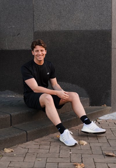 Figura masculina sentada en unos escalones de piedra, vistiendo una camiseta atlética negra y pantalones cortos negros, calcetines negros y zapatillas blancas. Cabello corto, sonriendo.