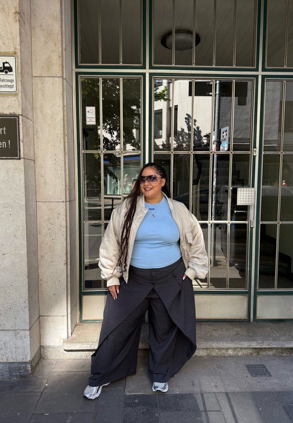 Femme aux longs cheveux tressés, portant des lunettes de soleil, une veste légère, une chemise bleue et un pantalon large et foncé, debout devant des portes vitrées sur le trottoir de la ville.