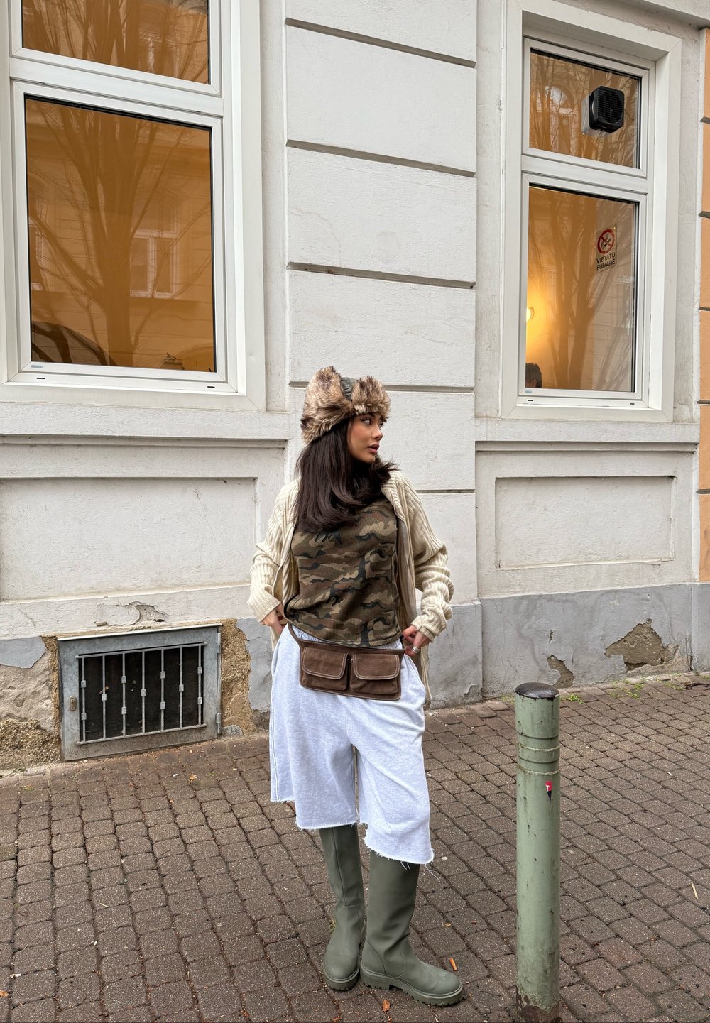 Tanktop mit Camouflage-Muster, cremefarbener Strickcardigan, weiße Shorts, grüne Gummistiefel und ein Hut mit Pelzbesatz. Braune Hüfttasche und Steinpflaster sind sichtbar.