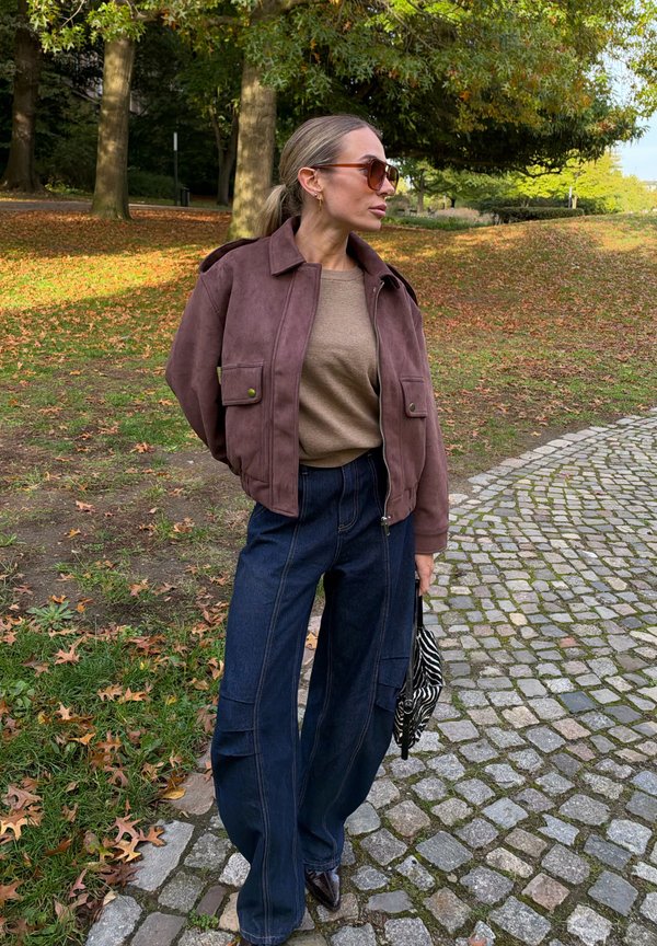 Una mujer con gafas de sol, una chaqueta marrón y vaqueros de pernera ancha está de pie en un camino empedrado de un parque otoñal y lleva un bolso estampado.