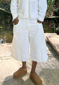 Witte katoenen oversized shorts met zijzakken, met een relaxte pasvorm. Bruine instapklompen van zacht materiaal. Natuurlijke buitenomgeving.