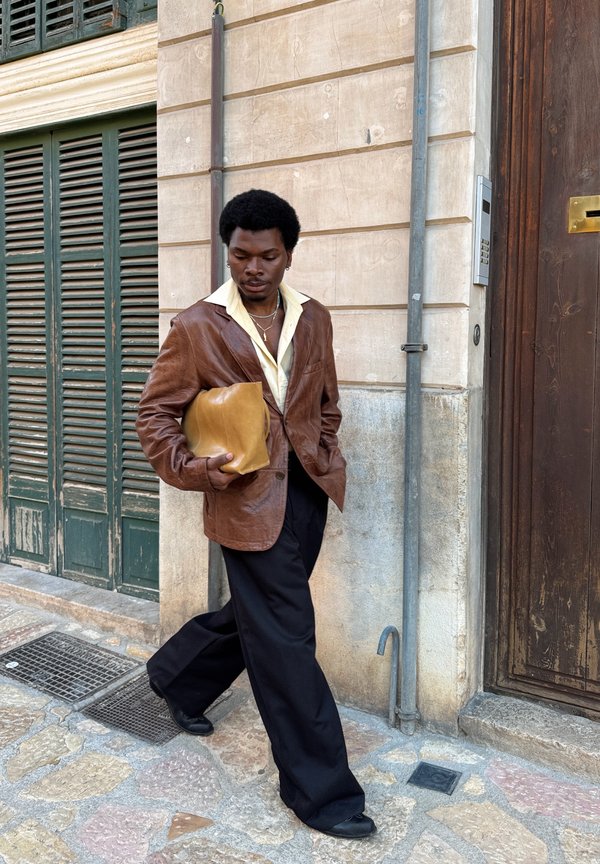 Blazer de couro castanho com uma camisa amarela por baixo, calças largas pretas, segurando uma clutch beje. Parede texturizada e persianas verdes ao fundo.