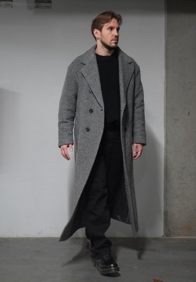 Abrigo largo de lana gris con solapas anchas, botones dobles, llevado sobre un suéter negro y pantalones negros. Zapatos negros completan el atuendo.