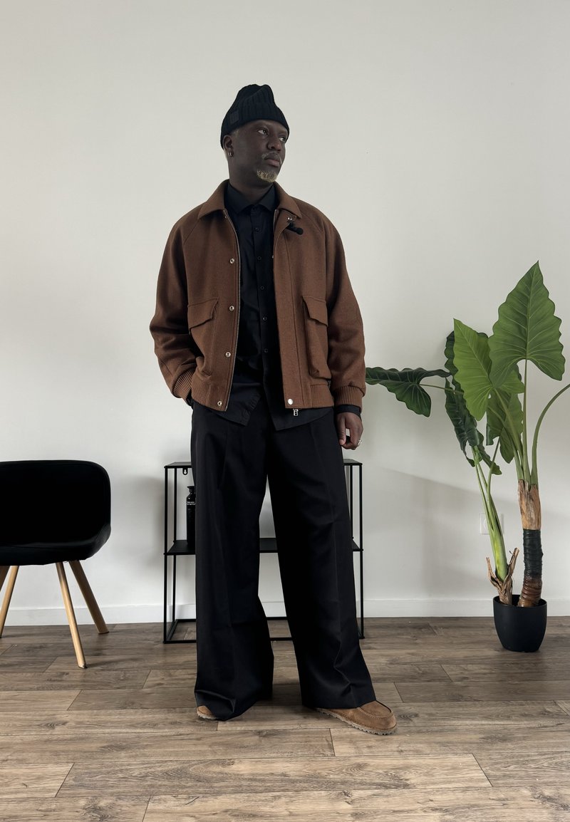 Hnědá vlněná bunda s dvěma předními kapsami, černá košile na knoflíky, černé široké kalhoty a hnědé boty. Zelená rostlina v pozadí.