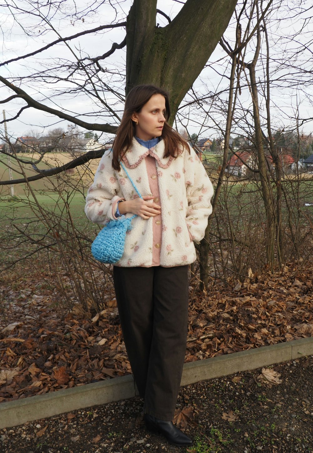 Veste en fausse fourrure à motif floral, teintes beige clair et rose. Sac tissé bleu, pantalon foncé et bottines noires. Décor naturel en plein air.