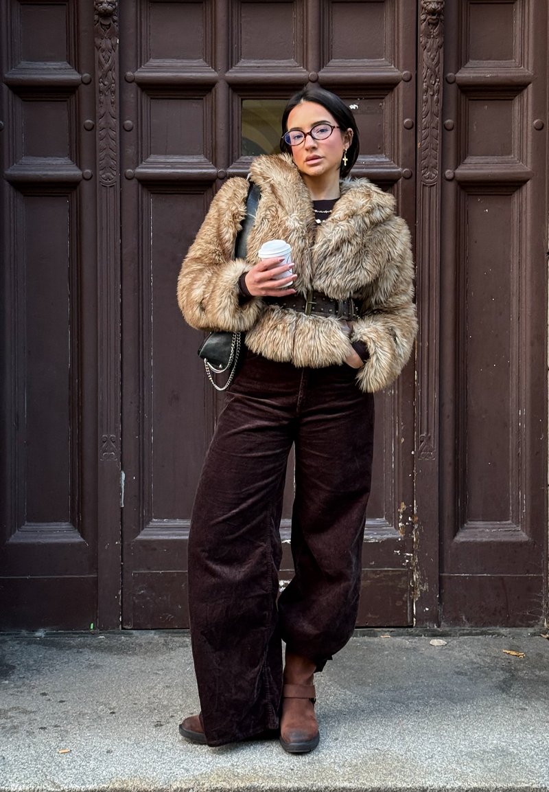 Casaco bege de pele sintética com gola, cinto preto; calças largas de bombazine castanho escuro; segurando uma chávena de café; mala preta de tiracolo.