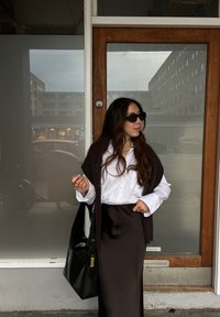 Geantă tote din piele neagră, cămașă albă cu butoni, un șal maro aruncat pe umeri, fustă lungă maro, păr ondulat, stând lângă o ușă de sticlă.